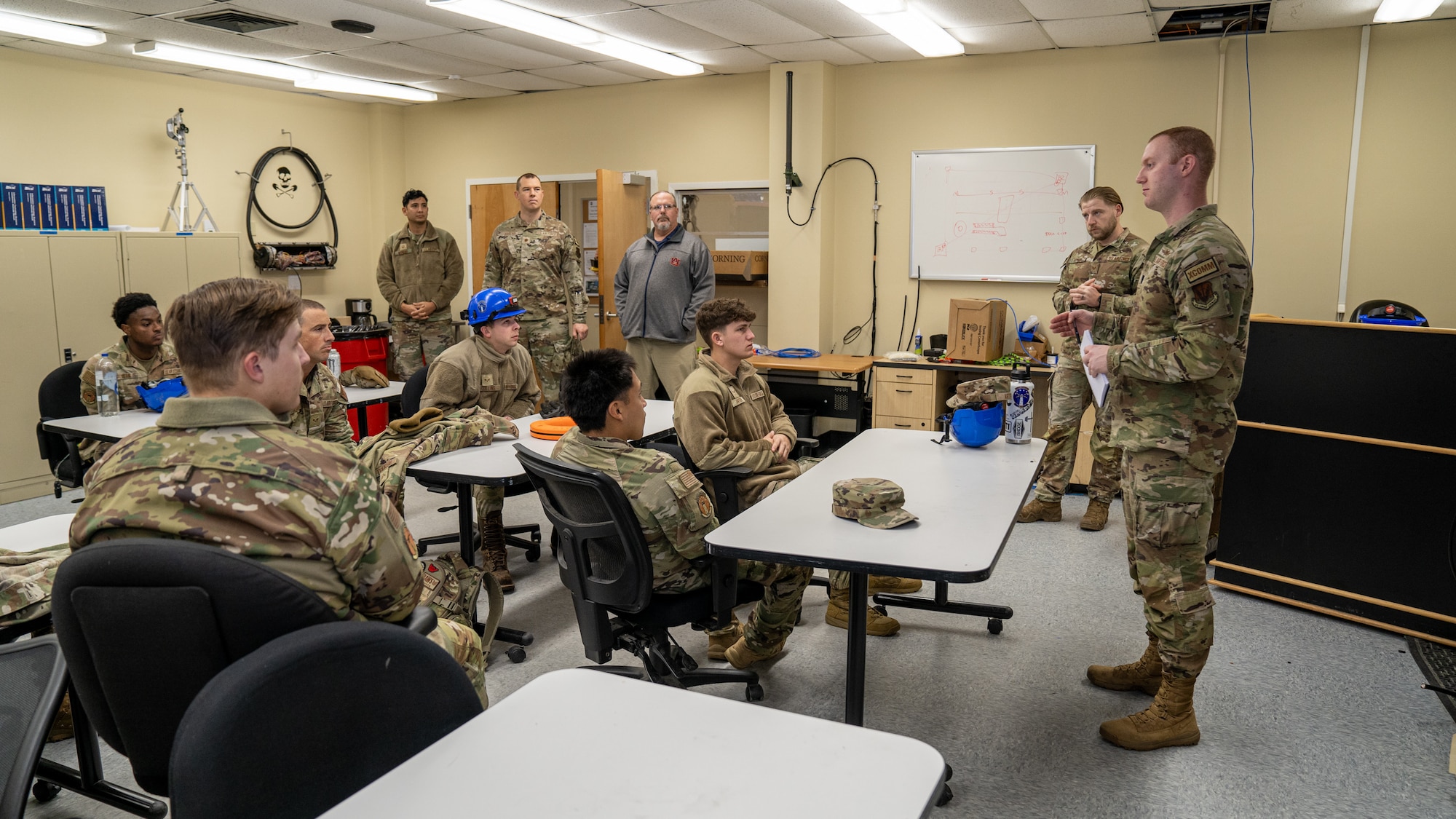 A serviceman in an operational camouflage pattern uniform briefs a room of about 10 service members.