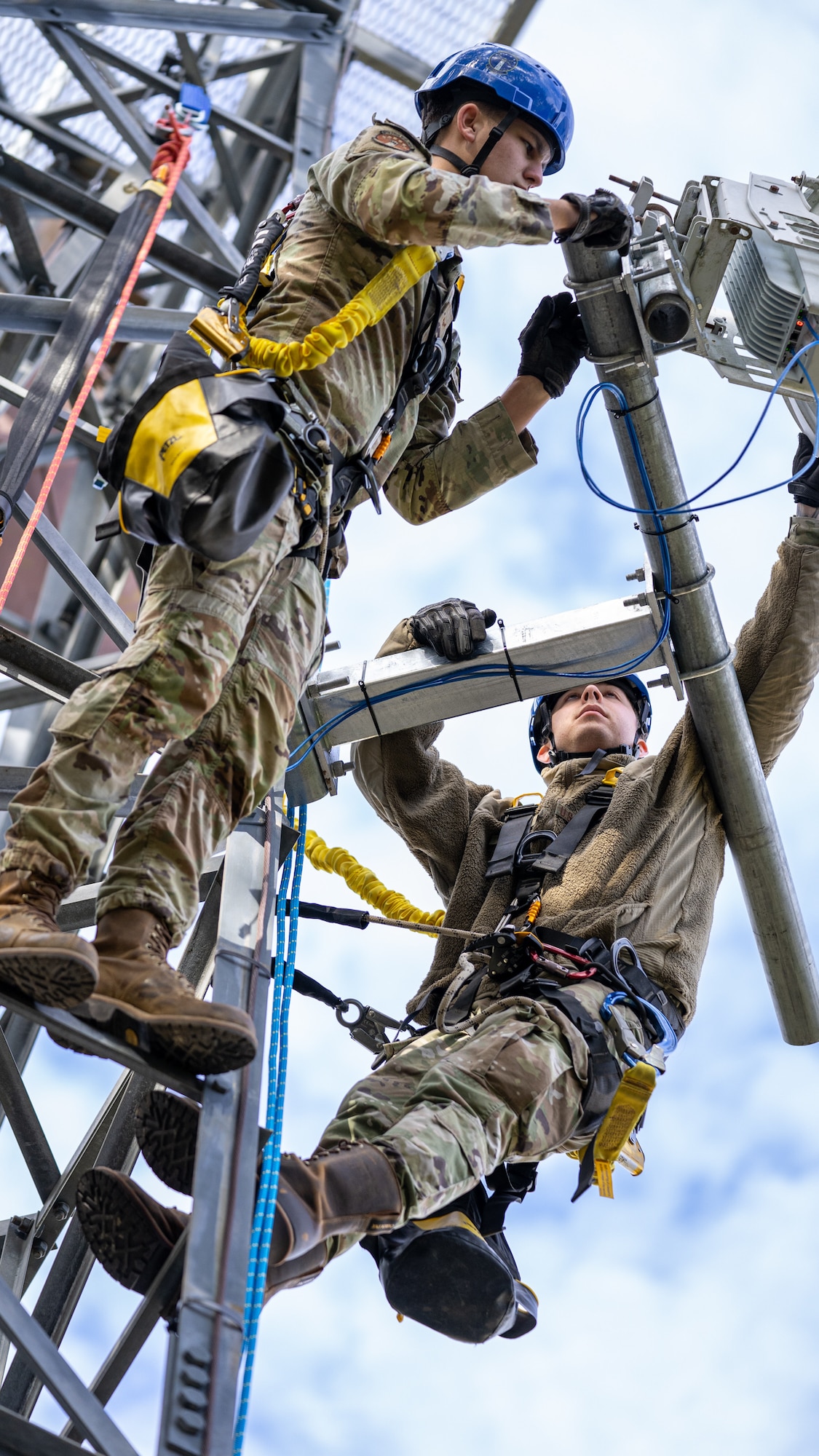 Two servicemen in operational camouflage pattern uniforms wearing tower climbing harnesses and hardhats adjust a disc antenna.