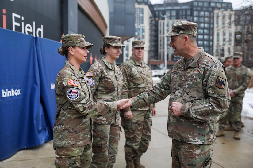 Two uniformed service members shake hands and smile while several others look on.