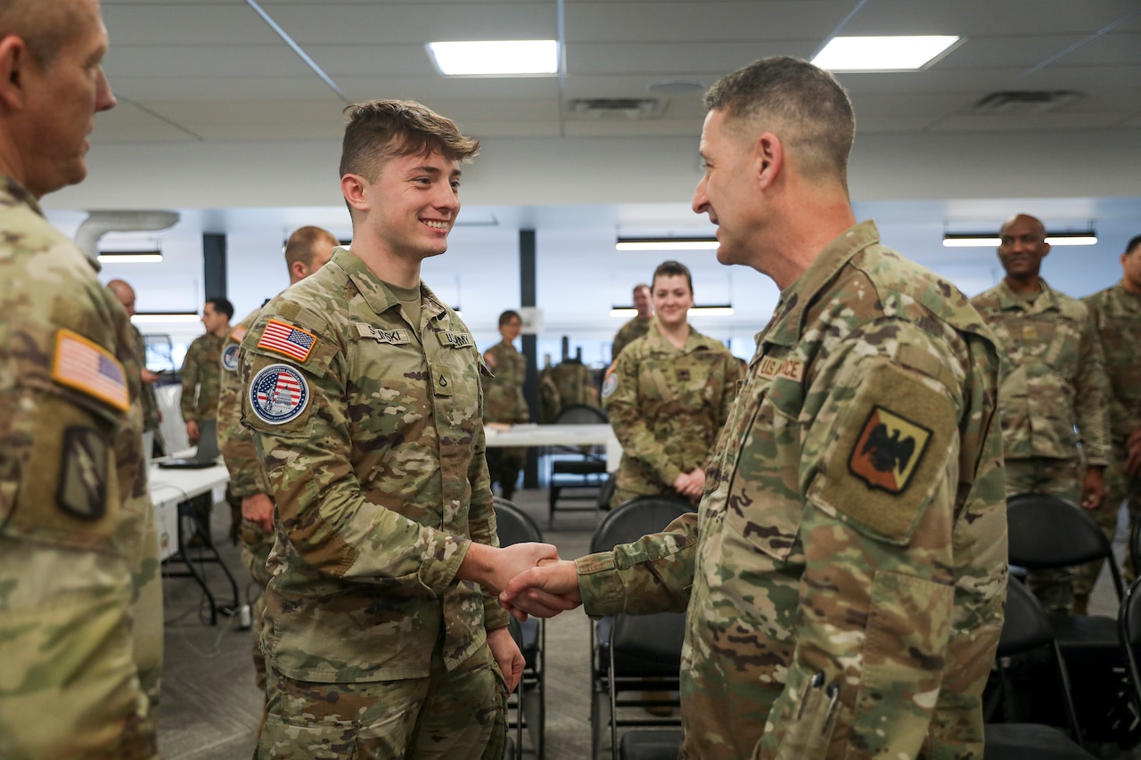 Two uniformed service members shake hands and smile while several others look on.