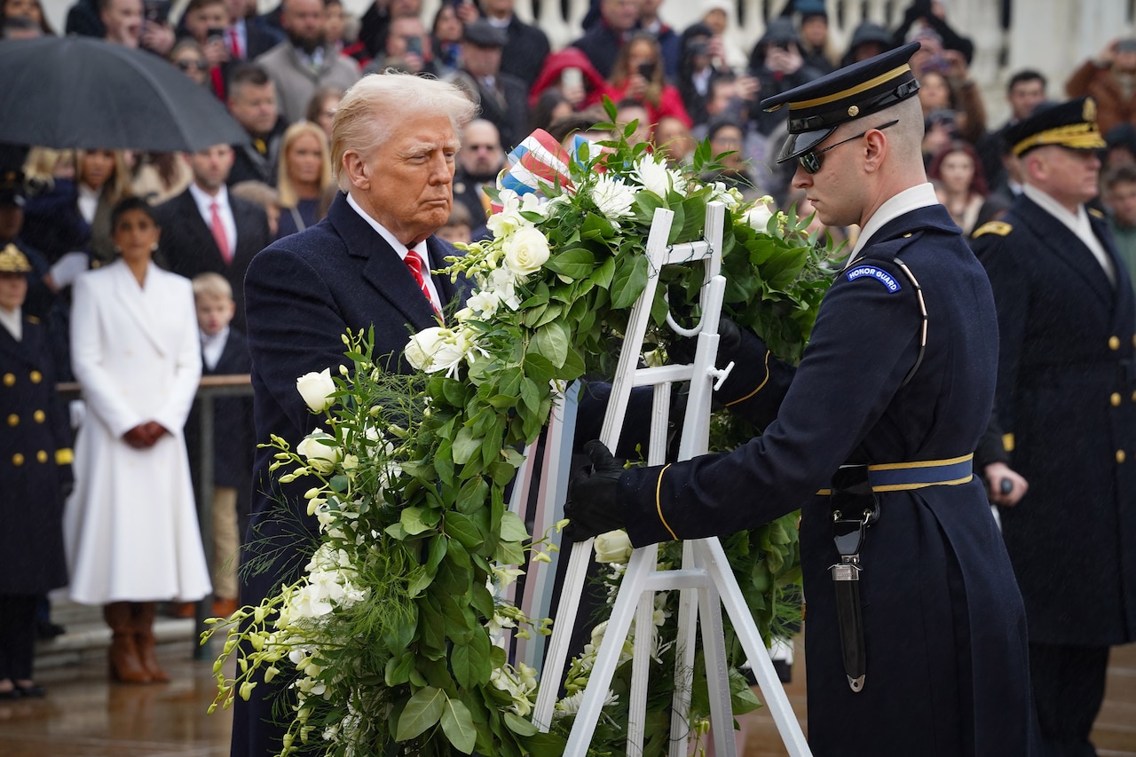 A man wearing business attire looks at a wreath that a uniformed service member is holding in front of a large crowd.