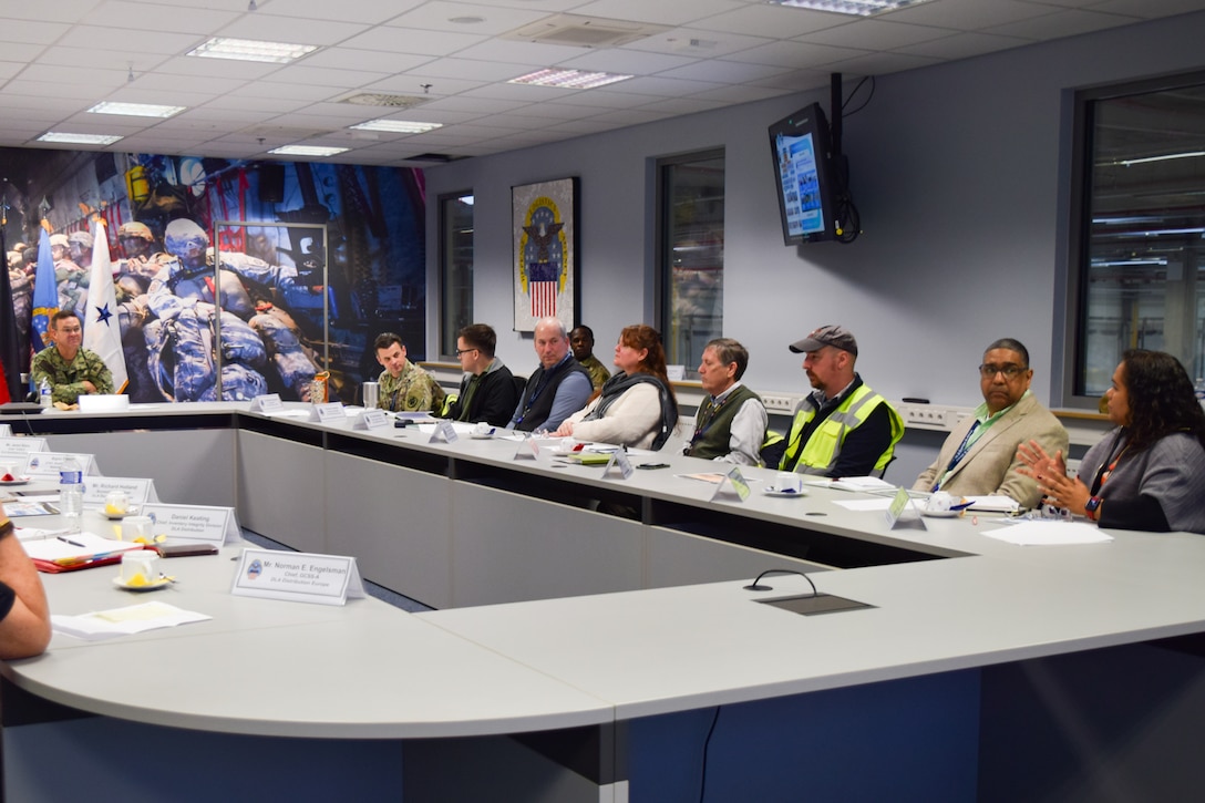 A group of people sit around a conference room table with a man wearing a camouflage military uniform sitting at the head of the table.