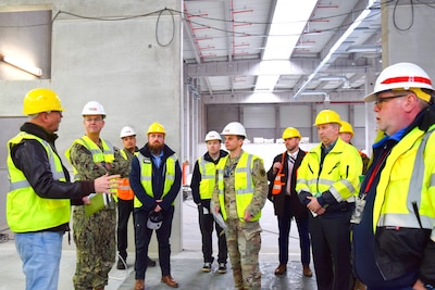 A group of people wearing reflective vests and hard hats stand around talking in an under-construction warehouse.