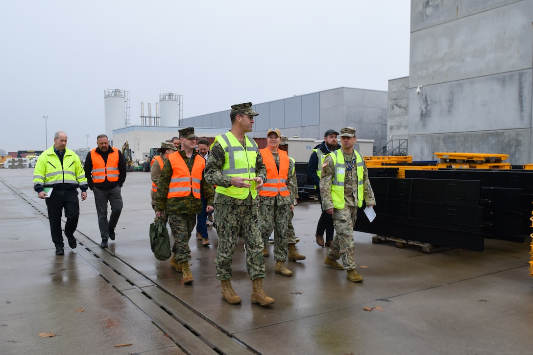A group of people in reflective vests walk outside past warehouses on an overcast day.