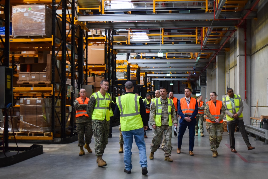 A group of people wearing reflective vests walk through a warehouse.