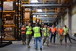 A group of people wearing reflective vests walk through a warehouse.