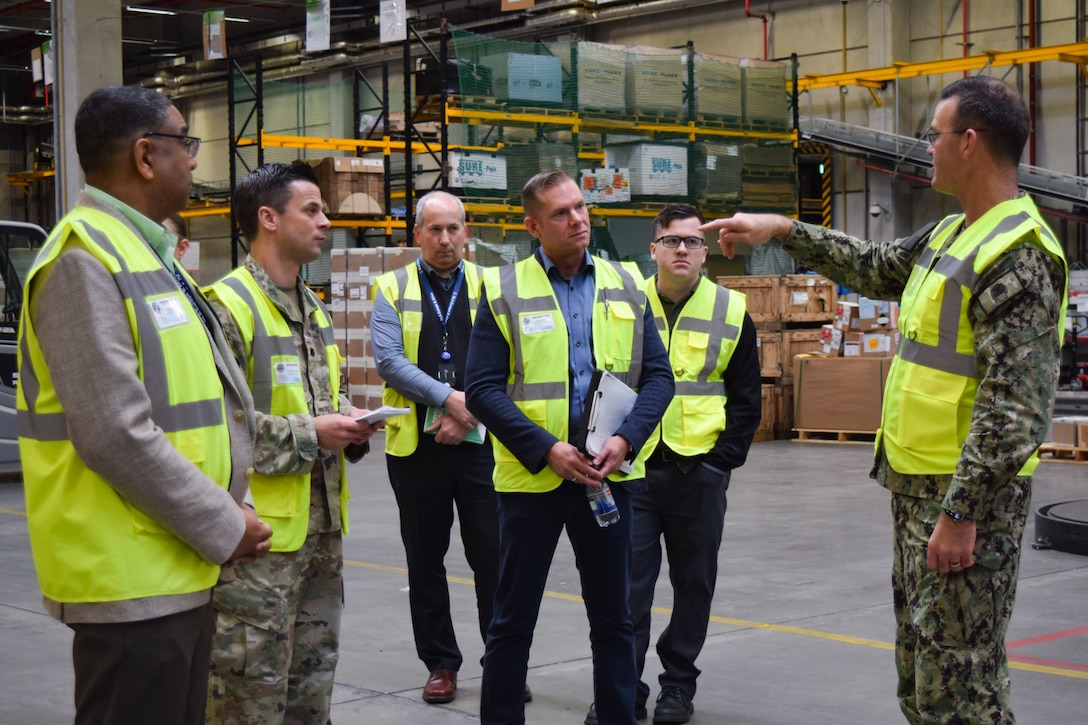 A man wearing a camouflage military uniform and reflective vest speaks to a group of people wearing reflective vests in a warehouse.