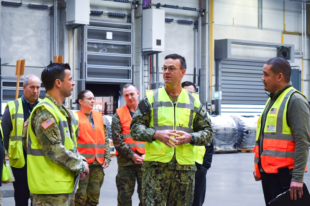 A group of people in reflective safety vests speak to one another in a warehouse.