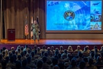 A military General speaks on stage to audience members in a theater.