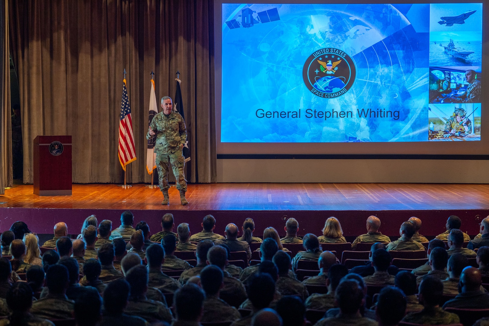 A military General speaks on stage to audience members in a theater.