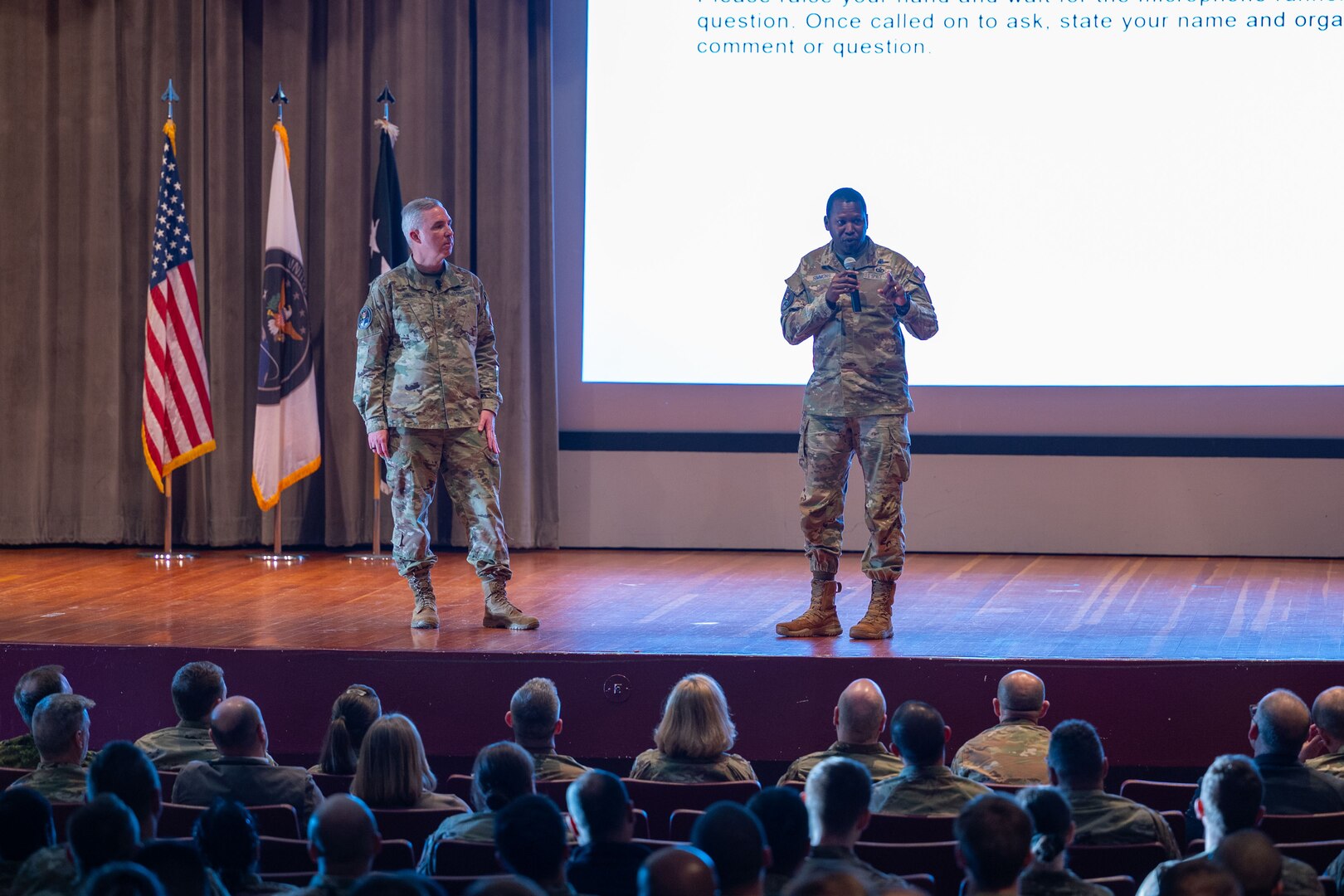 Two military leaders speak on stage to an audience in a theater.