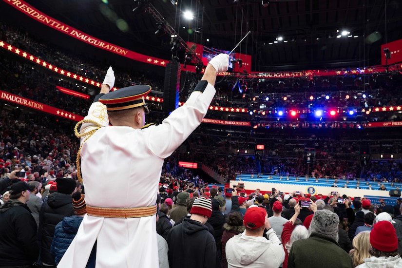 A conductor, in ceremonial dress surrounded by hundreds of people in an arena, signals to a band during a performance under red, white and blue lights.