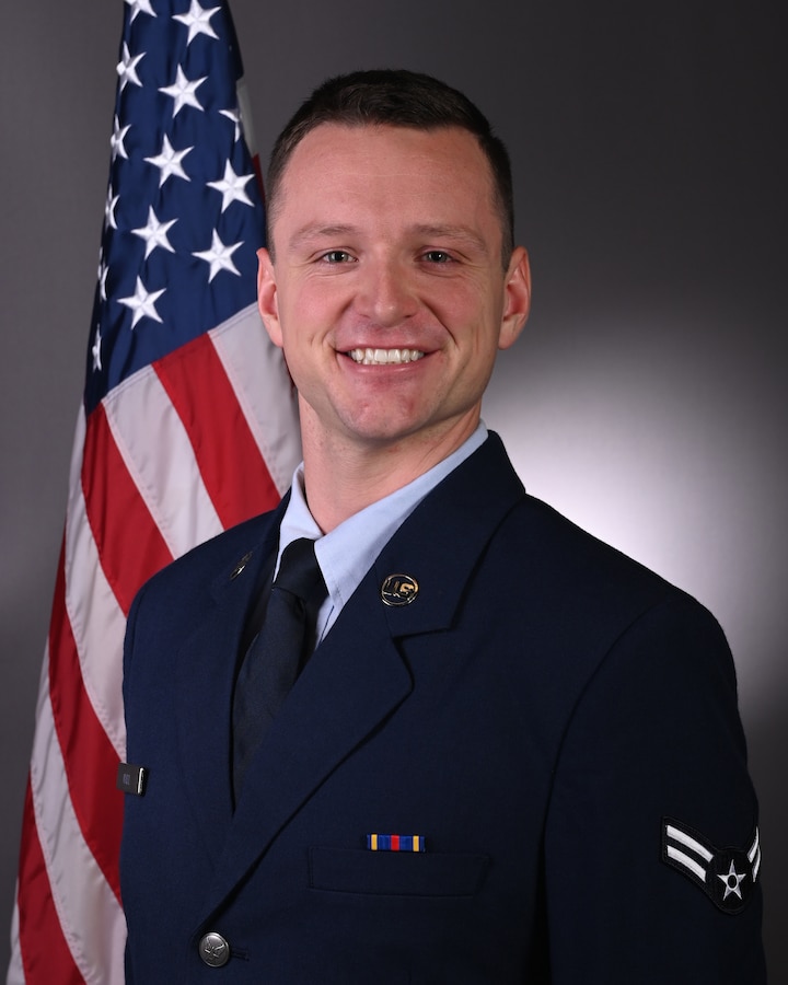 Official photo of A1C Christopher Hall in front of the American flag. He is wearing his blue service dress uniform