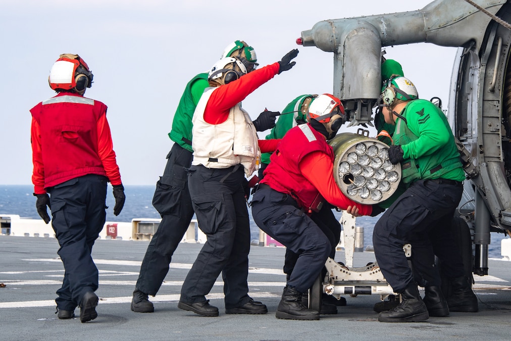 Sailors in green and red shirts load a weapon onto a partially visible helicopter aboard a ship at sea.