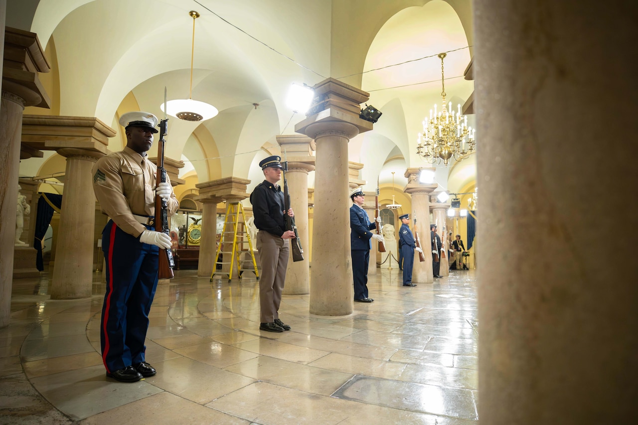 A group of service members practices standing and holding ceremonial weapons inside an elegant hall.