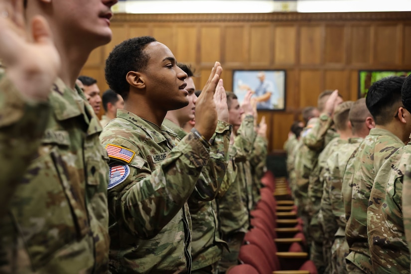 A group of soldiers stand in an audience lifting one hand in the air to pledge.