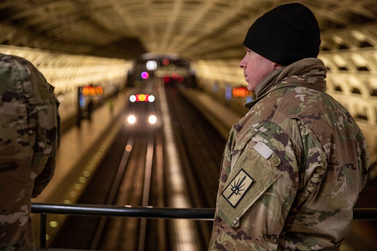 A soldier stands watch in an underground metro station as a train pulls in next to a platform below.