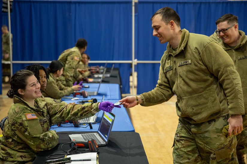 A soldier sitting on a table in a gymnasium passes a card to another soldier standing at the other side.