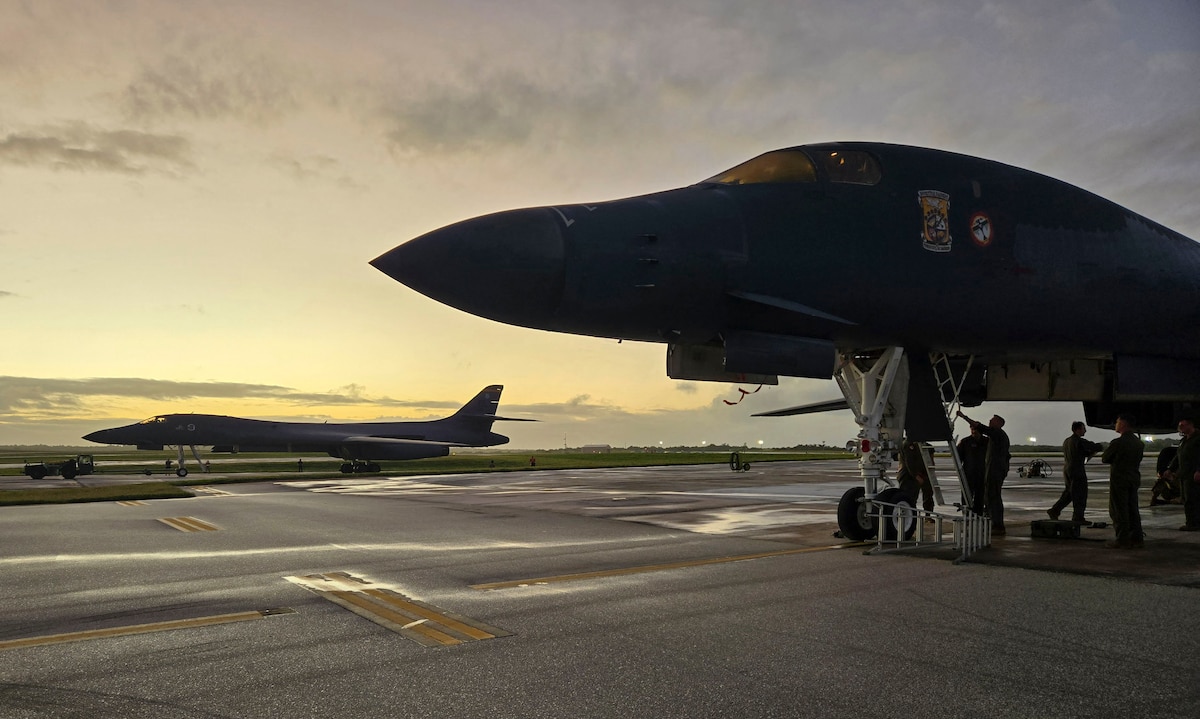 Two Air Force bomber aircraft are parked on a wide and open tarmac with the sun setting in the background. Two men work on the underside of the closest plane.