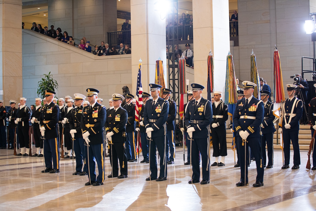 More than a dozen service members from multiple branches stand in formation in a large room where civilians observe them from a stairway.