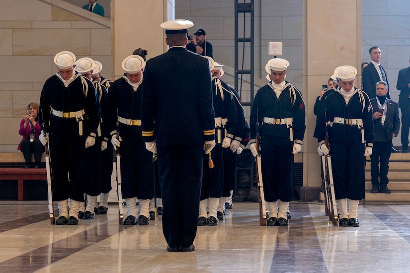 Sailors in dress uniforms stand in formation with heads bowed as a fellow sailor stands in front of them looking at them.