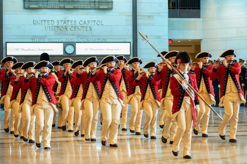 Army musicians in Revolutionary War-era uniforms perform in formation in a room, led by a drum major.