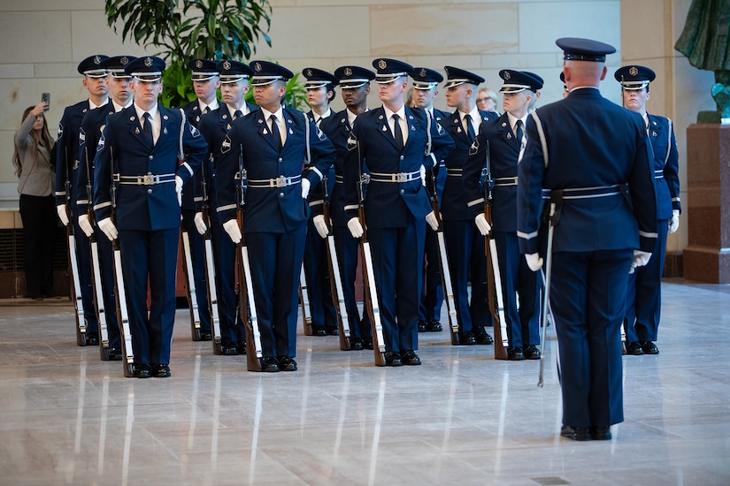 More than a dozen guardians in dress uniforms stand in formation as an fellow guardian faces them in a room.