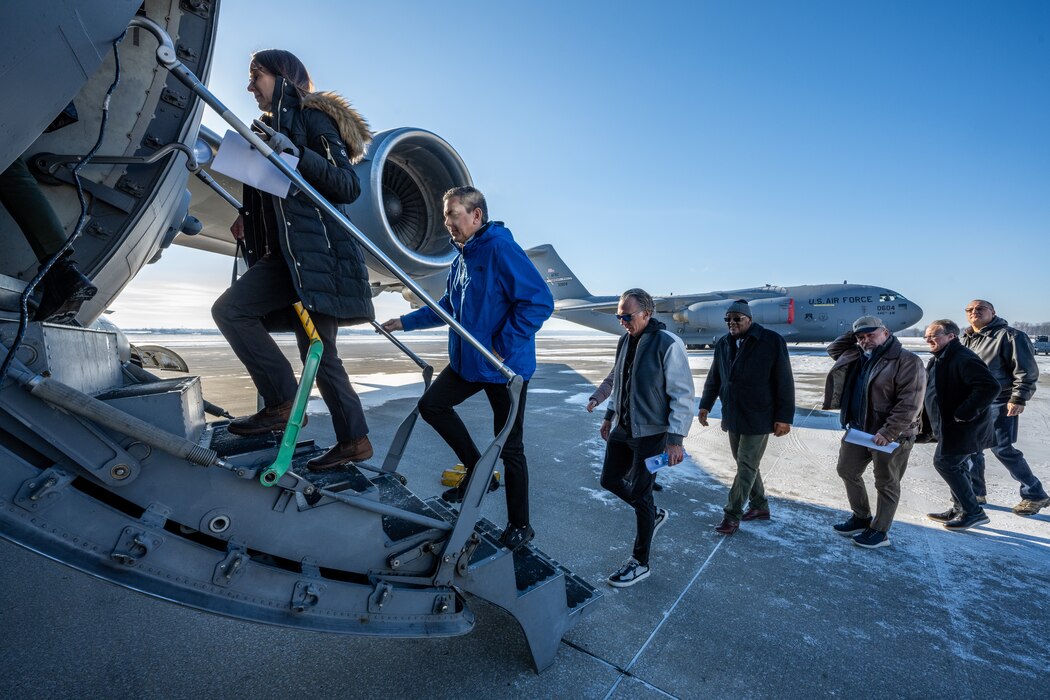 Two civilians in winter coats walk up a small aircraft staircase while a line of others follow. A large grey aircraft sits in the distance.