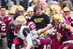 A man with a headset and who wears a beanie cap is seen on the sidelines of a football game with football players rushing in front of him.