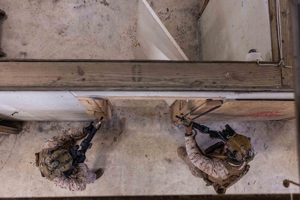 Two Marines in tactical gear hold weapons before entering the door of an open structure as seen from above.