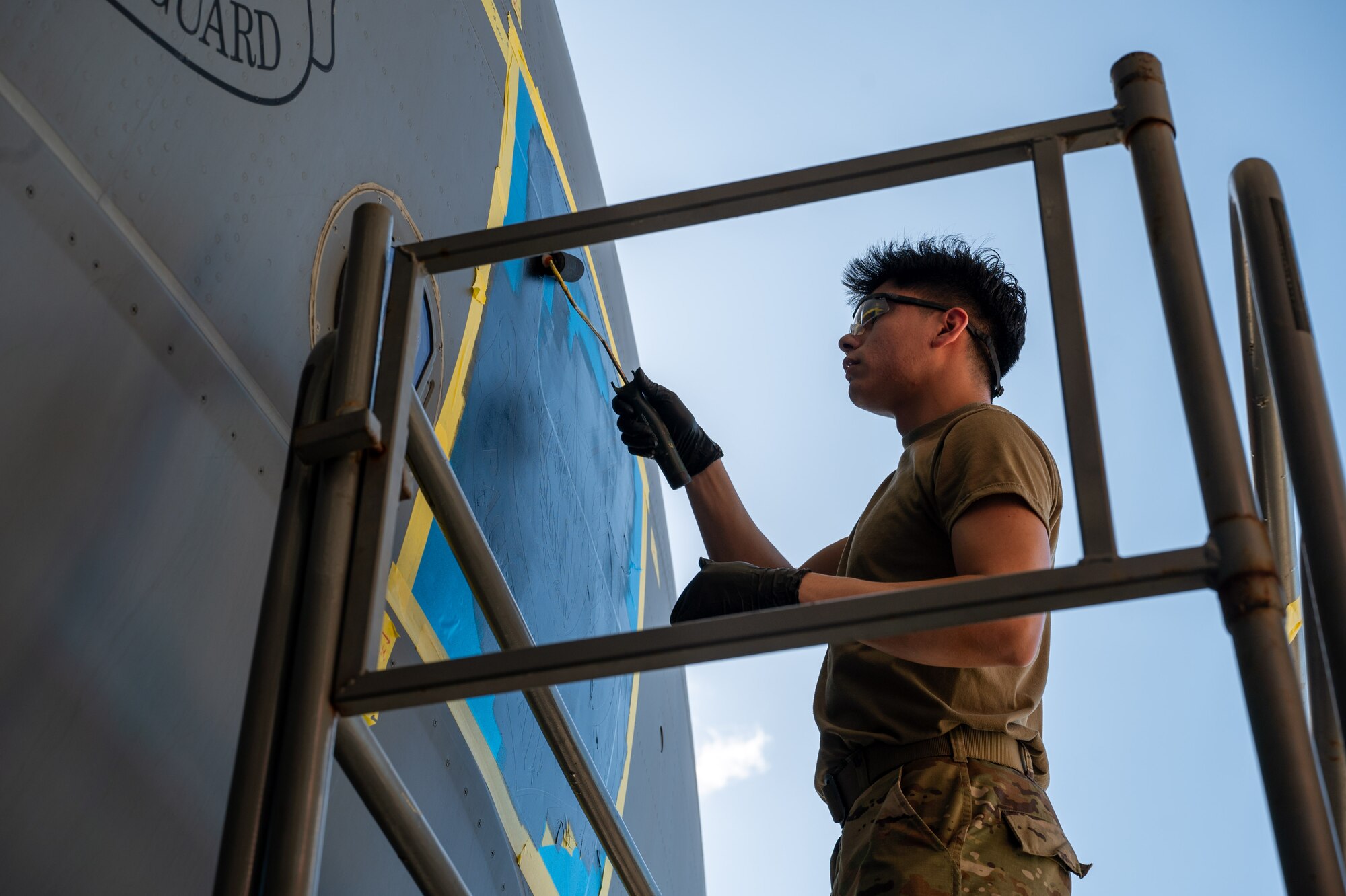 An Airman wearing black gloves uses a paint roller on a large blue square located on a C-17 Globemaster III aircraft.