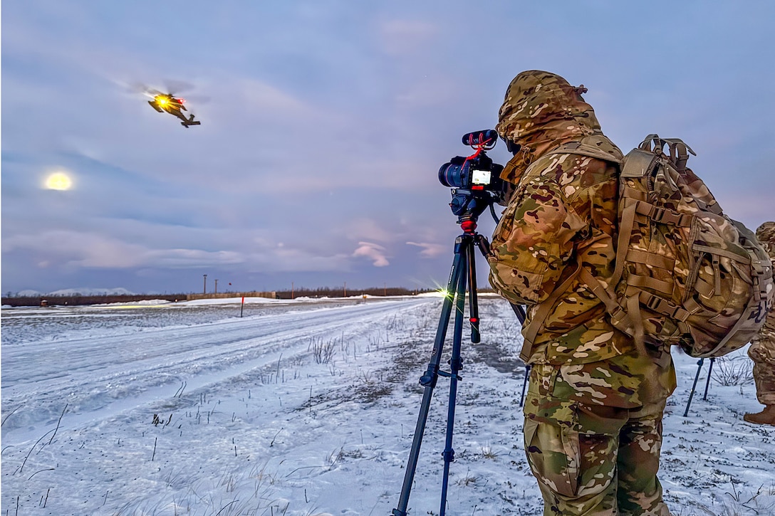 A soldier in winter gear stands behind a tripod-mounted camera toward a helicopter flying over a snowy field.