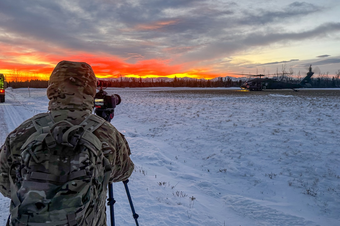 A soldier in winter gear looks through the lens of a tripod-mountain camera on a snowy field toward an orange-streaked sky.