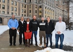 Six men and one woman stand outside of a large office building. the ground is covered in snow.