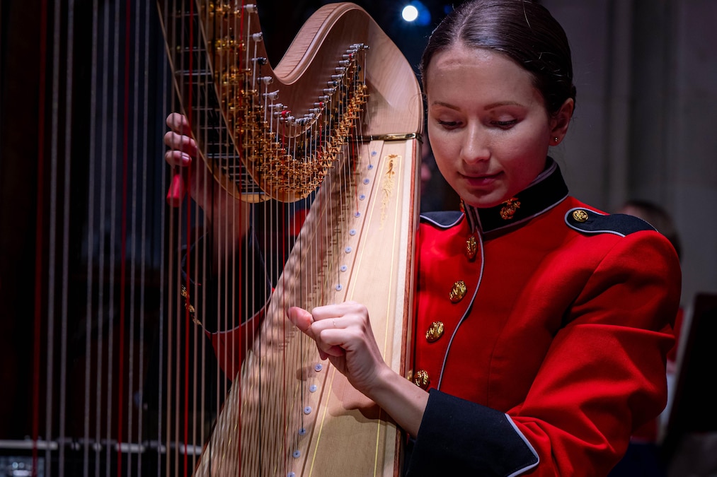 A Marine in ceremonial dress tunes a harp as a spotlight shines in the background.