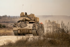Two heavily armored vehicles move through a hilly dry terrain.