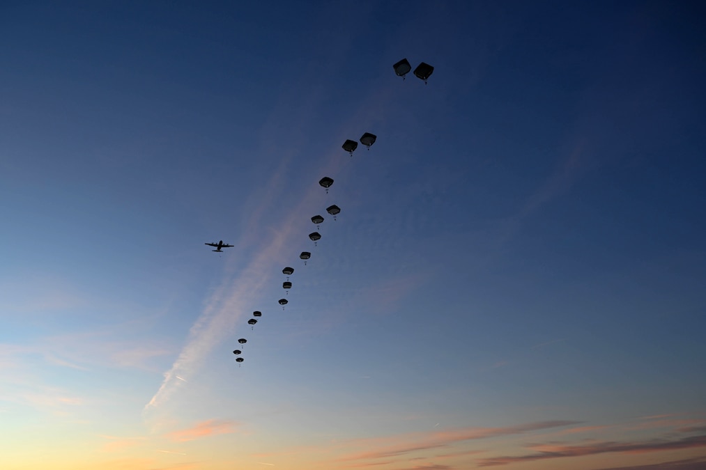 A line of silhouetted paratroopers descend from a military aircraft against a dusky blue sky with hints of orange in the horizon.