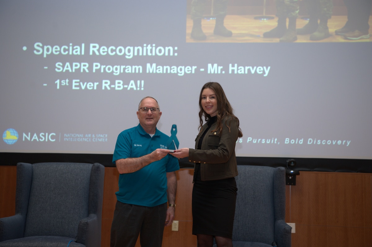 One female posed for a photo holding an award with a male next to her. They are smiling and gathered together in a professional setting.
