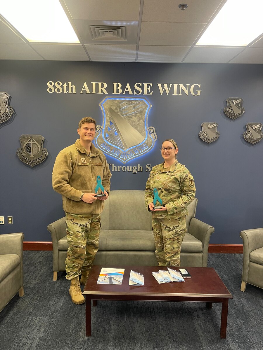One male and one female in an Operational Camouflage Pattern hold an award for a photo. They are smiling and gathered together in a professional setting.