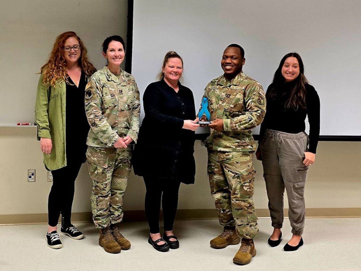 Five individuals pose for a photo, with one male in an Operational Camouflage Pattern uniform being awarded. A female in OCP also stands nearby, along with three other individuals dressed in civilian clothing. They are smiling and gathered together in a professional setting.