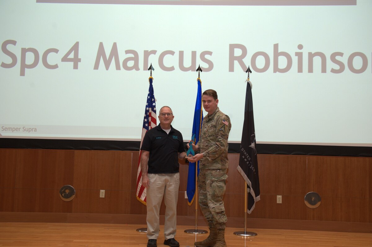 One male in an Operational Camouflage Pattern uniform being awarded by an individual dressed in civilian clothing. They are smiling and gathered together in a professional setting.