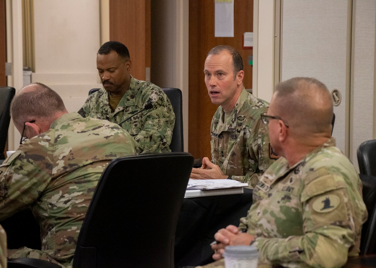 A man in a green military uniform speaks to a group of military personnel.