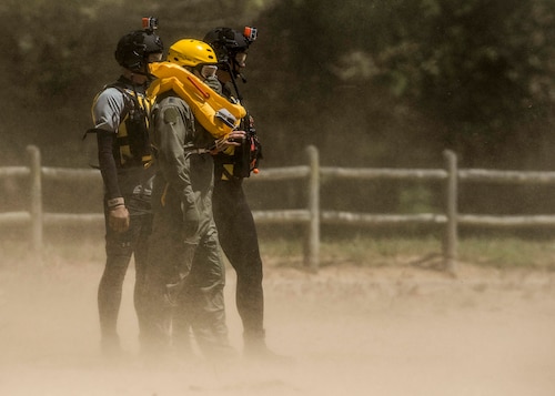 Survive, Evade, Resist, and Escape instructors from Scott Air Force Base and Lt. Col. Brandon Dow, 54th Airlift Squadron, stands in the dust kicked up by a UH-60 Blackhawk during Scott's first-ever water extraction training with the 6-101st Aviation Regiment of Fort Campbell, Ky., Lake Carlyle, Ill., June 7, 2017.  Dow, Lt. Col. Brooke Matson, 375th Operations Support Squadron, and Maj. Madison Basil Jr., Headquarters Air Mobility Command, received hoist, water survival, and flare training to learn how to survive in aquatic situations long enough to be rescued. The 101st Combat Aviation Brigade has served in almost every single military operation since the Vietnam War, including combat, peacekeeping, and humanitarian.  By partnering with Air Mobility Command’s SERE instructors, this training allows for joint-service coordination while preparing aerial crew members to survive in austere locations after being displaced from their aircraft.  (U.S. Air Force photo by Staff Sgt. Jodi Martinez) (U.S. Air Force photo by Staff Sgt. Jodi Martinez)