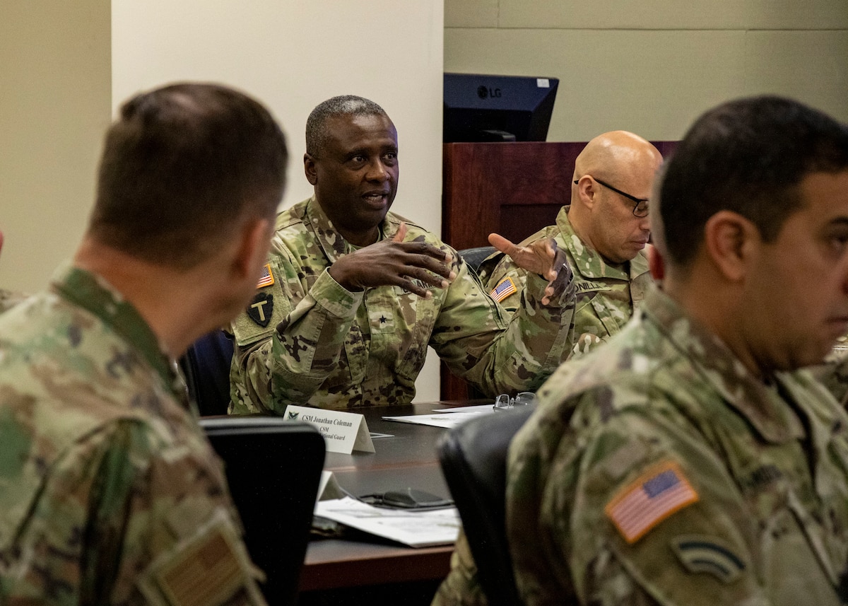 A man in a green military uniform speaks before a group of military personnel.