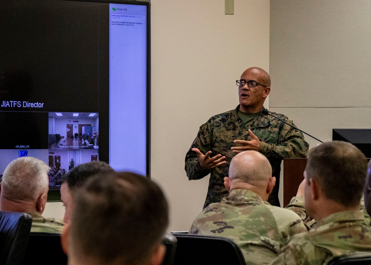 A man in a green military uniform speaks before a group of military personnel.