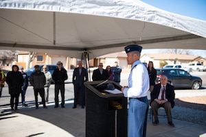 Military man in blues stands at podium to deliver speech under white tent while a crowd watches