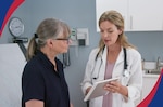 Female doctor. wearing white lab coat holds tablet in front of middle-aged patient sitting on an exam table.