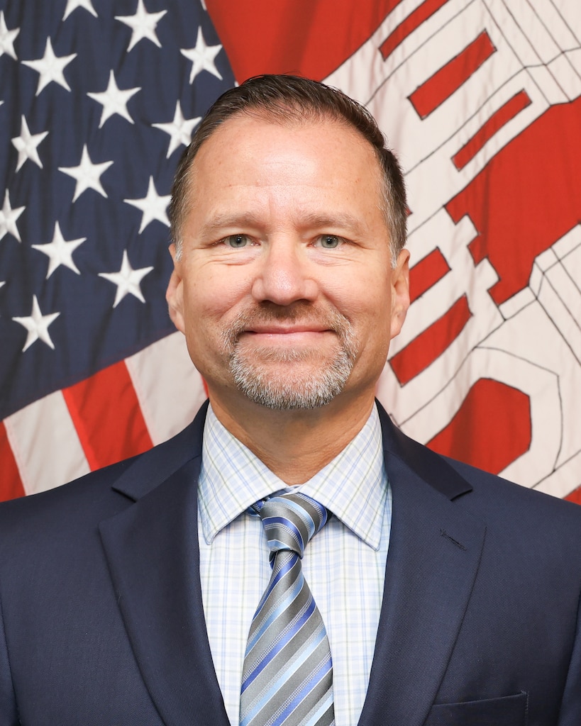 Man in a suit and tie standing in front of a US Flag and USACE flag