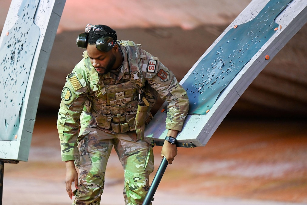 An airman wearing headphones picks up a target board filled with bullet holes.