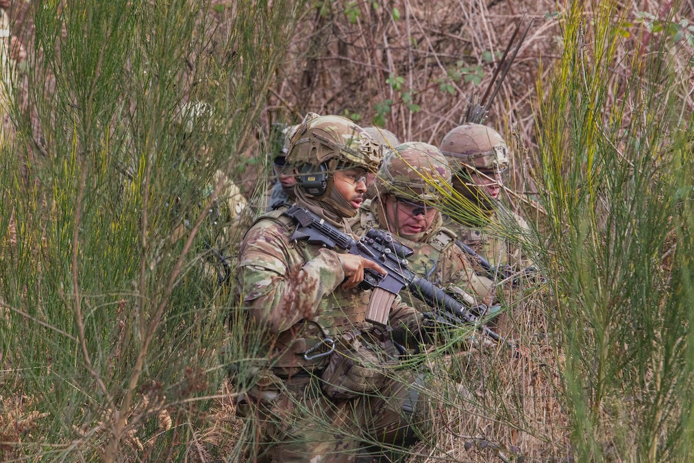 Six soldiers kneel among shrubs wearing combat gear and holding weapons during the day.